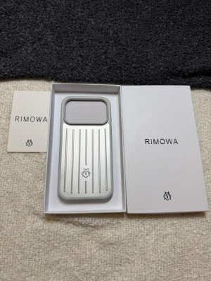 Rimowa silver mobilskal i aluminium - Snyggt mobilskal från Rimowa i silverfärgad aluminium med ikoniska räfflade linjer och logotyp på baksidan. Skalets design är stilren och robust med en rektangulär form och stora utskärningar för kamera och knappar. Perfekt för dig som vill ha ett exklusivt och tåligt skal.