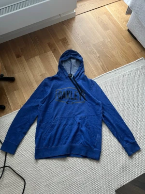 Blå hoodie från Oakley med logga - Snygg blå hoodie från Oakley med svart logga tryckt på bröstet. Klassisk känguruficka framtill och justerbar huva med svarta snören. Regular fit och mjukt material som känns skönt mot huden. Perfekt för dig som gillar sportig och avslappnad stil.