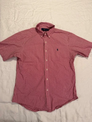 Rutig röd skjorta från Ralph Lauren - Kortärmad skjorta från Ralph Lauren i rödvitt rutigt mönster. Skjortan har button-down krage, broderad logga på bröstet och klassisk slim fit passform. Tillverkad i mjuk bomull som känns skön mot huden. Perfekt för en avslappnad men ändå stilren look.