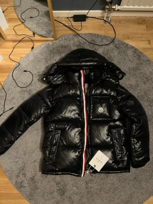 Svart Moncler dunjacka med huva - Snygg svart dunjacka från Moncler med glansig finish, huva och ikonisk logga på bröstet. Jackan har dragkedja med röd, vit och blå detalj samt två stora fickor framtill. Perfekt för kalla dagar och riktigt stilren look. NFC tagg funkar så klart 