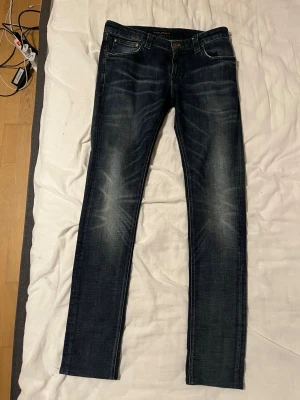 Mörkblå Nudie Jeans 30/34 straight fit - Säljer ett par mörkblå jeans från Nudie Jeans i storlek 30/34. Jeansen har en klassisk straight passform med snygga slitningar och kontrastsömmar. Tillverkade i bomull och perfekta för dig som gillar en tidlös look.