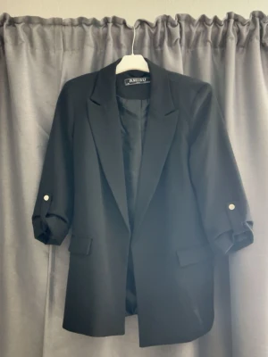 Svart kavaj från Ambiu med knappar - Stilren svart kavaj från Ambiu med klassisk krage och uppvikta ärmar som har dekorativa knappar. Kavajen har två fickor framtill och en rak passform. Perfekt för en trendig och snygg look.