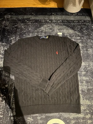 Svart kabelstickad tröja Polo Ralph Lauren - Kabelstickad svart tröja från Polo Ralph Lauren med klassisk röd broderad logga på bröstet. Tröjan har rund halsringning, långa ärmar och ribbstickade muddar vid ärmslut och nederkant. Perfekt för en stilren och avslappnad look.