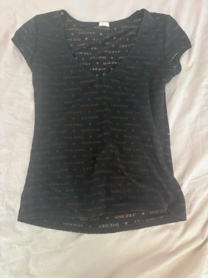 Svart topp med logotyp från Diesel - Svart topp från Diesel med korta ärmar och v-ringning. Tunt material med diskret mönster av Diesel-loggor över hela plagget. Snygg och enkel passform som funkar till många stilar.