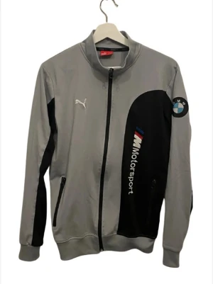 Grå och svart Puma BMW kofta  - Säljer en grå och svart Puma BMW Motorsport kofta i storlek S. Jackan har dragkedja framtill, två fickor med dragkedja och broderade loggor från både Puma och BMW. Snygg sportig look med detaljer i blått och rött på bröstet.