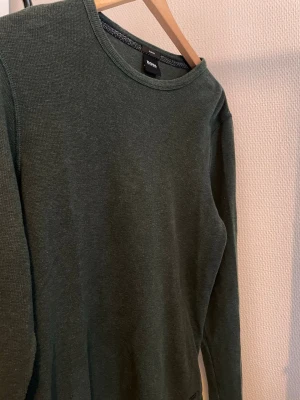 Mörkgrön stickad tröja från Hugo Boss - Snygg mörkgrön stickad tröja från Hugo Boss i slim fit-modell. Tröjan har rund halsringning och är tillverkad i ett mjukt, ribbat material som ger en clean och stilren look. Perfekt för dig som gillar minimalistisk stil.