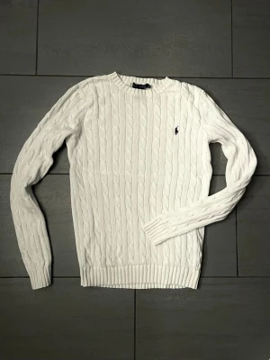 Vit kabelstickad tröja Polo Ralph Lauren - Kabelstickad vit tröja från Polo Ralph Lauren med rund halsringning och den klassiska broderade loggan på bröstet. Tröjan har ribbade muddar vid ärmslut och nederkant. Perfekt för dig som gillar en clean och tidlös stil.