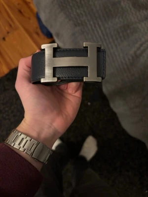 Mörkblått H läderbälte med H-spänne - Snyggt mörkblått bälte med ett H i äkta läder med det ikoniska H-spännet i borstat metall. Perfekt accessoar för att lyfta din outfit och ge en exklusiv touch. Passar till jeans eller kostymbyxor och är ett måste för dig som gillar stilrena detaljer. Anbänt 1-2 gånger