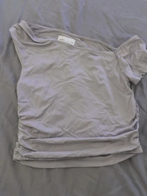 Ljusgrå offshoulder topp från Lefties - Trendig ljusgrå offshoulder topp från Lefties köpt i Spanien, mjukt material. Toppen har en asymmetrisk design med en ärm och passar perfekt till jeans eller kjol för en snygg look. 