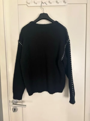 Svart stickad tröja med vita detaljer -  svart stickad tröja med vita sömdetaljer längs axlar och ärmar. Tröjan har en avslappnad passform, rund halsringning och långa ärmar. Perfekt för dig som gillar enkel men snygg stil.
