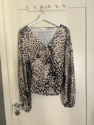 Leopardmönstrad blus med v-ringning - Trendig blus med leopardmönster i beige och svart. Djup v-ringning och långa ärmar med resår vid handlederna. Snygg omlott-effekt framtill och lätt glansigt material som ger en lyxig känsla. Perfekt för att lyfta din outfit. (Finare på i verkligheten än på bild) 
