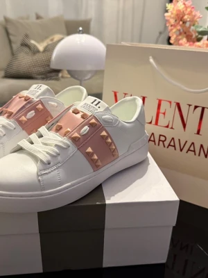 Valentino Rockstud sneakers vit/rosa - Snygga vita Valentino Rockstud sneakers med rosa band och ikoniska nitar i guld. Skorna har snörning och är tillverkade i skinn med rund tå och platt sula. Perfekta för dig som vill ha en exklusiv och trendig look.