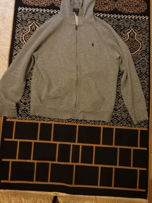 Grå zip hoodie från Polo Ralph Lauren - Säljer en klassisk grå zip hoodie från Polo Ralph Lauren med liten mörkblå logga på bröstet. Hoodien har dragkedja, två fickor fram och en skön huva. Tillverkad i mjukt sweatshirtmaterial, sopren och enkel att matcha till allt.