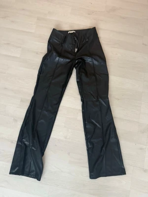 Svarta skinnbyxor med dragkedja - Säljer ett par svarta skinnbyxor med rak passform och silverfärgad dragkedja framtill. Byxorna har bälteshällor och en stilren look som passar till många olika outfits.