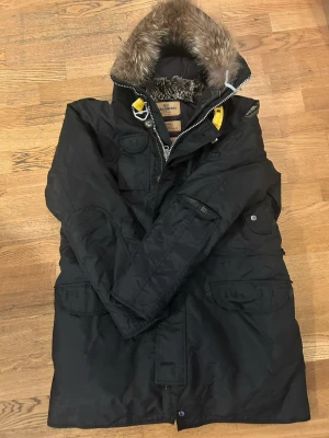 Svart Parajumpers parkas med fuskpäls - Svart parkajacka från Parajumpers med avtagbar fuskpäls på huvan, flera praktiska fickor och gul detalj vid halsen. Klassisk patch på ärmen och dragkedja framtill. Perfekt för kalla dagar och riktigt snygg streetstil.