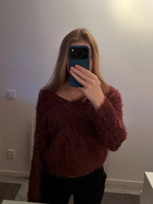 Vinröd fluffig stickad tröja - Vinröd glittrig stickad tröja med fluffig textur och bred urringning. Tröjan har långa ärmar och en snygg rosé detalj i ryggen med öppen slits och knytband. 