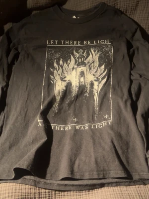 Svart långärmad t-shirt med tryck - Svart långärmad t-shirt med ett stort vitt tryck på framsidan som föreställer en kyrka i lågor och texten 'LET THERE BE LIGHT AND THERE WAS LIGHT'. T-shirten har en avslappnad passform och är tillverkad i mjuk bomull.