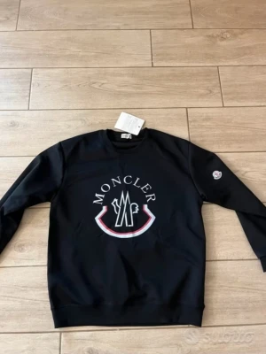 Svart sweatshirt från Moncler - Svart sweatshirt från Moncler med stort tryckt logomärke på bröstet och en liten logotyp på ena ärmen. Tröjan har rund halsringning och långa ärmar. Perfekt för en stilren och sportig look.