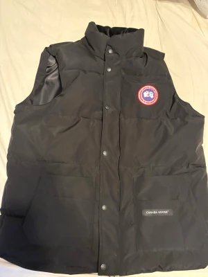 Svart dunväst från Canada Goose - Snygg svart dunväst från Canada Goose med hög krage, två stora framfickor och klassisk logotyp på bröstet. Västen har tryckknappar framtill och är vadderad för extra värme. Perfekt för lager-på-lager och streetwear-vibe.