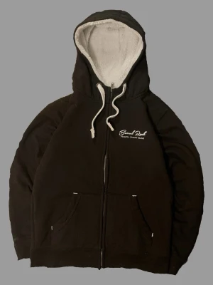 Svart hoodie Barrel Rock med dragkedja - Svart hoodie från Barrel Rock med vit text på bröstet och dragkedja framtill. Insidan är fodrad med mjuk, vit teddy och huvan har vita snören. Två fickor framtill och klassisk passform. Perfekt för dig som gillar en mysig och stilren look.