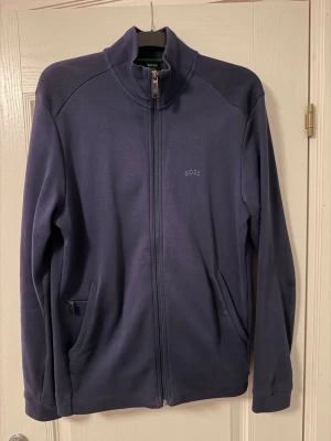 Mörkblå tröja från BOSS - Snygg mörkblå ziptröja från BOSS med hög krage och diskret logga på bröstet. Tröjan har två fickor med dragkedja och är tillverkad i ett mjukt material som passar perfekt till en avslappnad stil. Långärmad och enkel att matcha med jeans eller joggers.