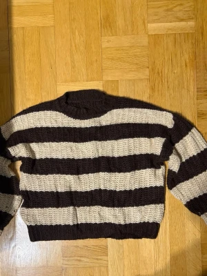 Brun- och beige randig stickad tröja - Säljer en chunky stickad tröja med breda bruna och beige ränder. Tröjan har en avslappnad passform, rund halsringning och långa ärmar. Perfekt för dig som gillar en retro och cozy vibe. Materialet känns mjukt och tjockt, perfekt för kyligare dagar.