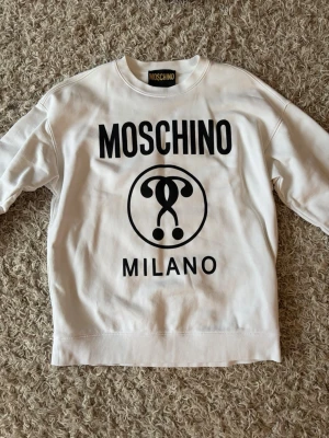 Vit sweatshirt från Moschino - Säljer en vit sweatshirt från Moschino med stor svart logga och texten 'MOSCHINO MILANO' på framsidan. Tröjan har rund halsringning och är tillverkad i mjukt bomullsmaterial. Perfekt för dig som gillar streetwear och vill sticka ut.