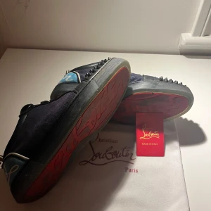 Christian Louboutin sneakers med nitar - Snygga svarta sneakers från Christian Louboutin med klassisk röd sula och coola svarta nitar på tån. Skorna har svarta snören, detaljer i blått och vitt samt en mix av skinn och mocka. Perfekta för dig som vill sticka ut med exklusiv streetstyle. Utan BOX! Säljer billigt för att bli av med