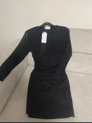 Svart prickig klänning från sisters point - Svart kortklänning från  sisters point med vita prickar och långärmade ärmar. Klänningen har rund hals och är gjord i ett tunt, transparent syntetmaterial som ger en trendig look. Perfekt för dig som gillar stilrena detaljer och mönster.