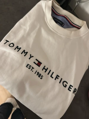 Vit sweatshirt Tommy Hilfiger M - Snygg vit sweatshirt från Tommy Hilfiger med klassisk logga och texten 'EST. 1985' på bröstet. Rund halsringning med ribbad kant och diskreta ränder på insidan av kragen. Perfekt för en clean och stilren look.💕