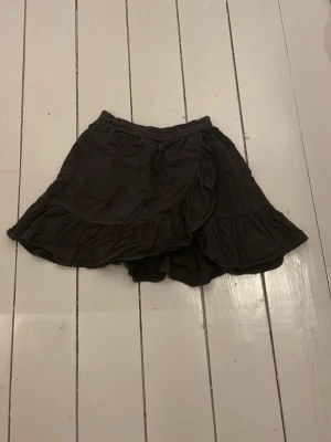 Svart volangkjol  - Kort svart omlottkjol med volangkant och elastisk midja. Kjolen har en flowy passform och är perfekt för en avslappnad men ändå trendig look.