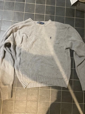 Grå stickad tröja från Polo Ralph Lauren - Klassisk grå stickad tröja från Polo Ralph Lauren med rund halsringning och diskret broderad logga på bröstet. Tröjan har ribbade muddar vid ärmslut och nederkant, perfekt för lager på lager och enkel att matcha till olika outfits. Finns en liten fläck på bröstet
