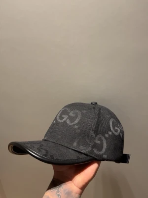 Svart keps från Gucci med GG-mönster - Snygg svart keps från Gucci med klassiskt GG-monogram över hela kepsen. Justerbar rem baktill i svart läder och böjd skärm med läderdetalj. Perfekt accessoar för en trendig streetwear-look.