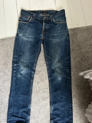 Blå jeans från Nudie Jeans Co - Säljer ett par klassiska blå jeans från Nudie Jeans Co med snyggt slitna detaljer och orange sömmar på bakfickorna. Modellen har slim fit, med fem fickor och knappgylf. Materialet är robust denim i bomull. Ett par riktigt fina jeans som jag säljer då de är lite för små för mig tyvärr.