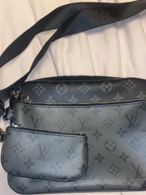 L V Svart axelväska  - Säljer en svart axelväska från L V med klassiskt monogrammönster. Väskan har två fack med dragkedja, justerbar axelrem och snygga silverdetaljer. Materialet är skinn och designen är stilren och modern, perfekt för dig som gillar exklusiva accessoarer.