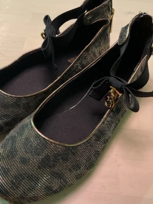 Glittriga ballerinaskor Michael Kors - Snygga ballerinaskor från Michael Kors i svart och guld med glittrigt leopardmönster. Rund tå, platt sula och svarta band med rosett och guldfärgade MK-detaljer vid ankeln. Perfekt för dig som gillar att sticka ut med coola detaljer och lyxig vibe.