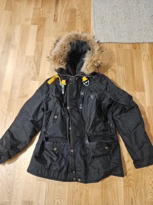 Svart parkajacka med päls - Svart parkajacka  med päls på huvan, coola gula detaljer och patch på ärmen. Jackan har flera fickor, dragkedja och knappar framtill. Insidan är fodrad med mjuk grå fleece. Perfekt för kalla vinterdagar och har en snygg, urban look. Large men sitter mer som en S enligt mig. Köptes här så vet ej om den är äkta eller ej. Därav priset.