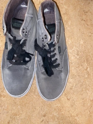 Grå höga sneakers från Vans - Säljer ett par grå höga sneakers från Vans i storlek 41. Skorna har svart snörning, rund tå och vit kant runt sulan. Ovandelen är i slitstark canvas med metallöglor och klassisk Vans-logga på sidan. Perfekt för dig som gillar streetstil.