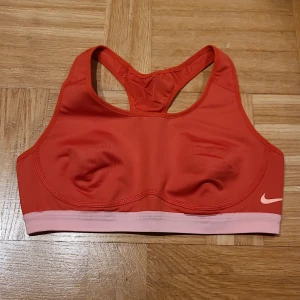 Röd sporttopp från Nike Dri-Fit - Nike Dri-Fit sporttopp i röd med ljusrosa kant nedtill. Modellen är ärmlös med brottarrygg och har ett litet Nike-logotryck på framsidan. Tillverkad i ett syntetmaterial som andas och håller dig torr under träningen. Perfekt för gymmet eller löprundan.