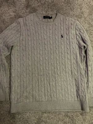 Grå stickad tröja från Polo Ralph Lauren - Säljer en grå stickad tröja från Polo Ralph Lauren med klassiskt kabelstickat mönster och rund hals. Tröjan har långärmade ribbade muddar och en broderad logga på bröstet. Perfekt för dig som gillar stilrena och tidlösa plagg.