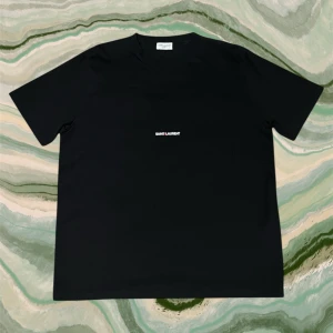 Svart Saint Laurent t-shirt - Svart t-shirt från Saint Laurent Paris med minimalistisk vit logga på tishan. Jag är 187 och den passar mig perfekt. Pris är ej hugget i sten. Skriv vid minsta lilla fråga😊