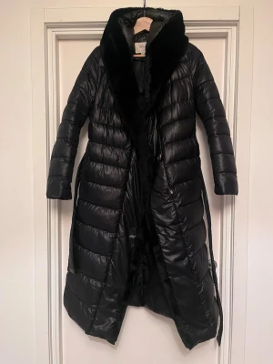 Svart lång pufferjacka från ZIAI - Trendig svart pufferjacka från ZIAI med krage i skönt teddymaterial, luva och bälte i midjan. Jackan är lång, har quiltad design och är perfekt för kalla dagar. Storlek S. Mycket bra skick! Upplagd på flera säljsidor, först till kvarn. 
