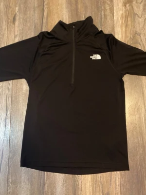 Svart half zip träningströja från The North Face - Säljer en svart tröja från The North Face med half zip och hög krage. Tröjan har korta ärmar och är tillverkad i ett syntetmaterial som är perfekt för träning eller aktiva dagar. Loggan är tryckt i vitt på bröstet.