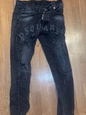 Svarta Dsquared2 ICON jeans - Säljer ett par svarta jeans från Dsquared2 med stor Dsquared-logga framtill och röd ICON-lapp bak. Jeansen har slitna detaljer, smal passform och klassisk femficksdesign. Materialet är jeans och färgen är mörkgrå/svart med coola tryck och distressed look.