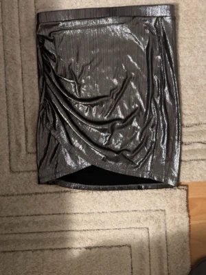 Glittrig silver kortkjol med omlott - Säljer en glittrig silverfärgad kortkjol med omlott-design och snygga draperingar framtill. Kjolen har ett skimrande tyg med subtila ränder och ger en riktigt cool vibe. Perfekt för dig som gillar att sticka ut med metallic och trendiga detaljer.