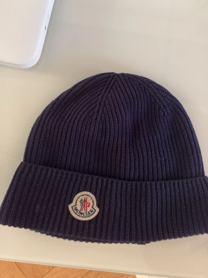 Mörkblå stickad Moncler mössa - Säljer en mörkblå ribbstickad mössa från Moncler med klassisk logotyp framtill. Mössan är tillverkad i mjukt material och har en bred uppvikt kant för extra stilpoäng. Perfekt för dig som vill ha en snygg accessoar till vintern. Den är 100% äkta 