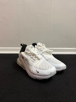 Nike Air Max 270 vita sneakers - Nike Air Max 270 sneakers i vit med svarta detaljer och synlig luftbubbla i hälen. Ovandelen är i ventilerande syntetmaterial med snörning och rund tå. Snygg och modern design som passar perfekt till streetwear och träning.