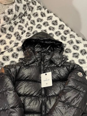 Moncler Maya  - Svart pufferjacka från Moncler i blankt syntetmaterial med huva och dragkedja. Jackan har Moncler-logga på ärmen och coolt serietidningsmotiv på insidan. Perfekt för dig som vill ha en varm och snygg jacka med streetkänsla.