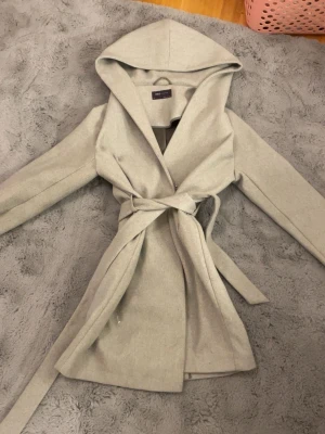 Grå kappa med huva och knytband - Snygg grå kappa med stor huva och knytband i midjan. Långärmad modell i mjukt material som ger en clean och stilren look. Perfekt för dig som gillar minimalistisk stil och vill ha något som känns både trendigt och bekvämt. Kan eventuellt tänka mig gå ner i pris