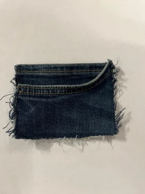 Blå jeans kuvertväska med fransar - Säljer en unik kuvertväska gjord av återvunnet blått jeansmaterial. Väskan har råa, fransiga kanter och en enkel ficka framtill. Perfekt för dig som gillar en cool och avslappnad stil. Ingen synlig logga eller märke.